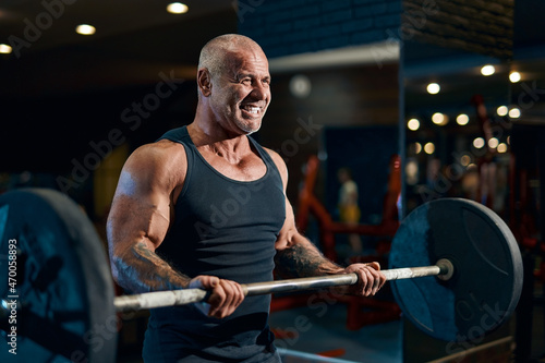  Emotional Older bald sportsman trains in the gym while pumping up biceps muscles with barbell