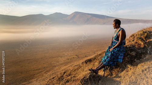 Masai Wwarrior sitting at the edge of one of Ngorongoro craters looking at the horizon and enjoying the surnrize photo