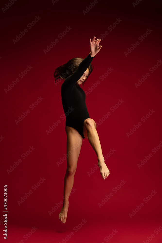 One emotional young flexible contemp dancer, ballerina jumping isolated on dark red background. Art, beauty, inspiration concept. One emotional young flexible contemp dancer, ballerina jumping isolated on dark red background. Art, beauty, inspiration concept.