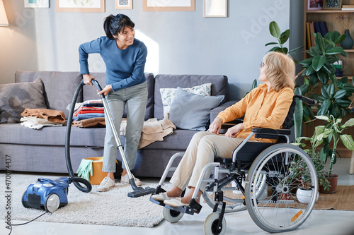 Housemaid using vacuum cleaner to clean the carpet in the room, she helping senior woman who using wheelchair