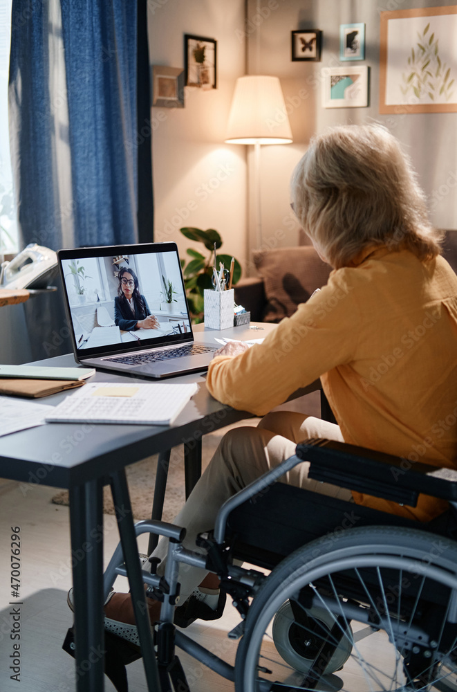 Rear view of senior woman sitting at the table in the room and using laptop for online conversation, she talking to businesswoman Rear view of senior woman sitting at the table in the room and using laptop for online conversation, she talking to businesswoman