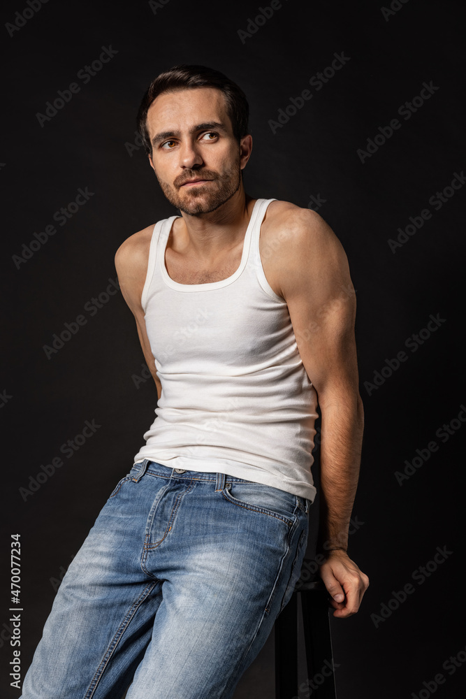 Classic portrait of young handsome bearded sad man in white undershirt and jeans isolated on dark background. Emotions, facial expression Classic portrait of young handsome bearded sad man in white undershirt and jeans isolated on dark background. Emotions, facial expression