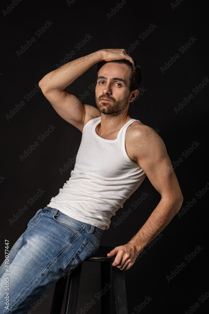 Classic portrait of young handsome bearded sad man in white undershirt and jeans isolated on dark background. Emotions, facial expression Classic portrait of young handsome bearded sad man in white undershirt and jeans isolated on dark background. Emotions, facial expression