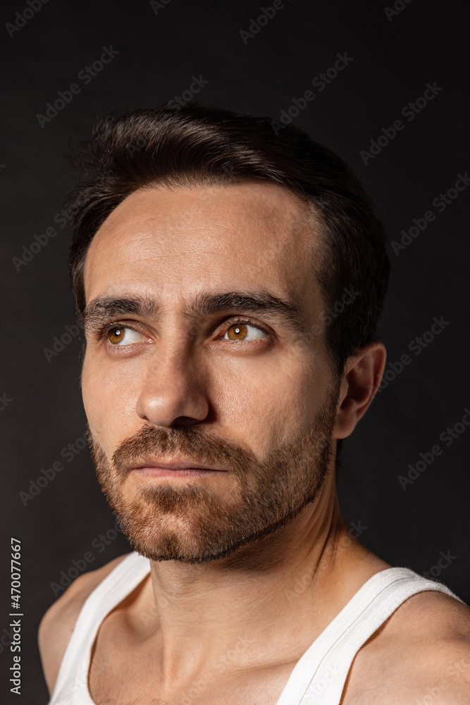 Obraz premium Close-up portrait of young handsome bearded man in white undershirt isolated on dark studio background. Emotions, facial expression