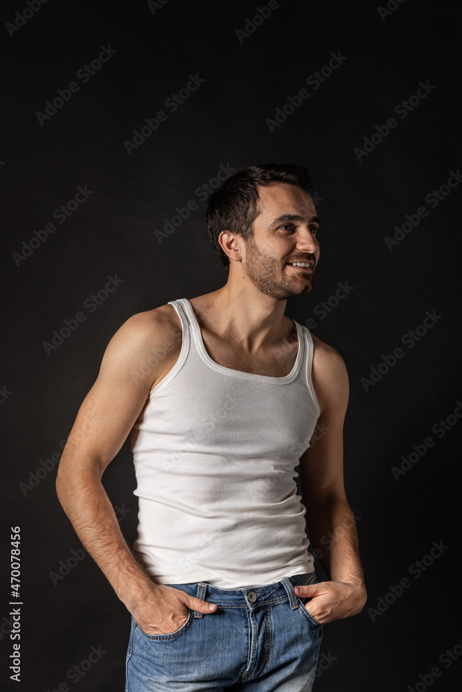 Portrait of young handsome bearded sad man in white undershirt and jeans posing isolated on dark background. Portrait of young handsome bearded sad man in white undershirt and jeans posing isolated on dark background.