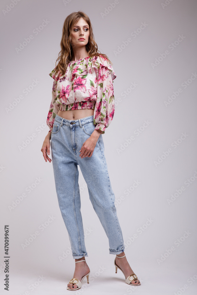 High fashion photo of a beautiful elegant young woman in a pretty blouse top with floral pattern in red, blue jeans posing over white, soft gray background. Slim figure. Studio Shot. High fashion photo of a beautiful elegant young woman in a pretty blouse top with floral pattern in red, blue jeans posing over white, soft gray background. Slim figure. Studio Shot.