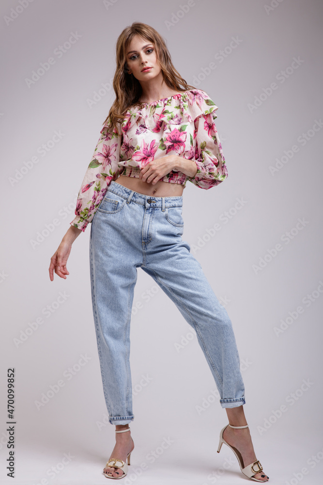 High fashion photo of a beautiful elegant young woman in a pretty blouse top with floral pattern in red, blue jeans posing over white, soft gray background. Slim figure. Studio Shot. High fashion photo of a beautiful elegant young woman in a pretty blouse top with floral pattern in red, blue jeans posing over white, soft gray background. Slim figure. Studio Shot.