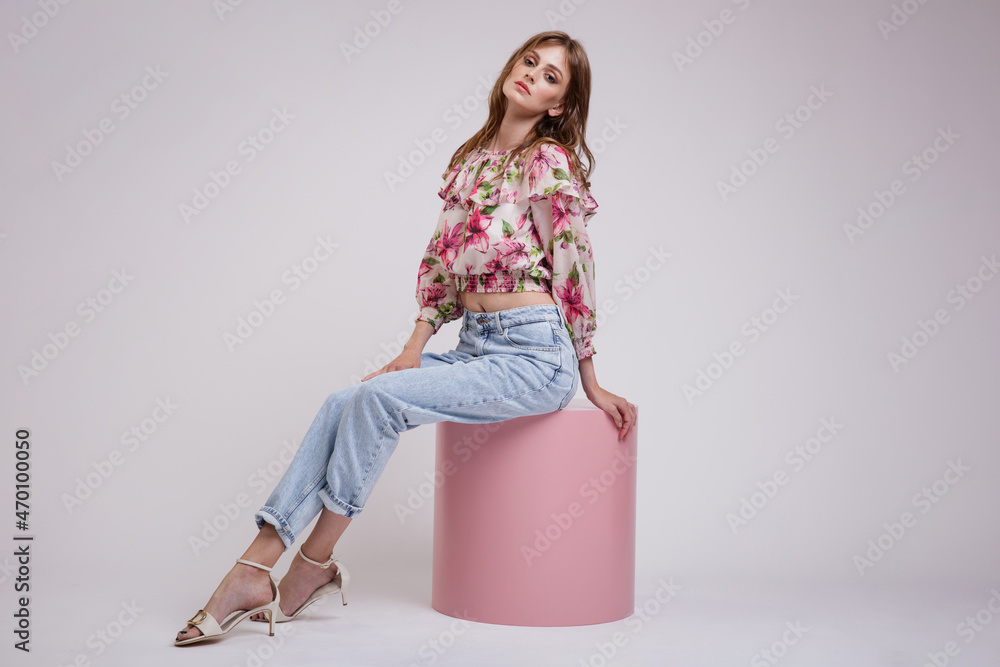 High fashion photo of a beautiful elegant young woman in a pretty blouse top with floral pattern in red, blue jeans posing over white, soft gray background. Model is sitting on a pink cube cylinder High fashion photo of a beautiful elegant young woman in a pretty blouse top with floral pattern in red, blue jeans posing over white, soft gray background. Model is sitting on a pink cube cylinder
