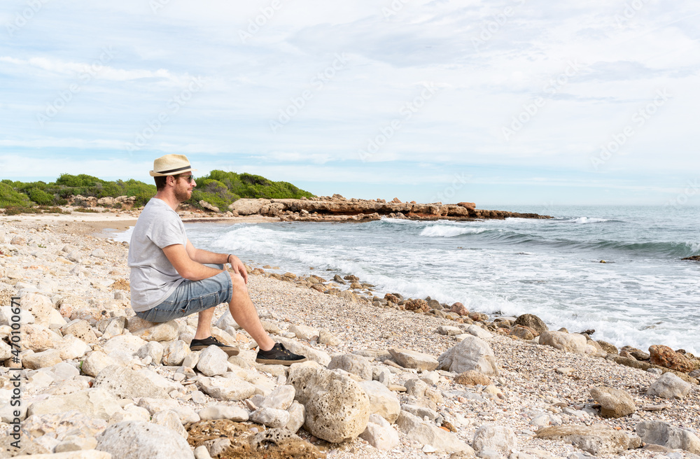 Side view of a smiling man sitting on a rock at a idyllic mediterranean beach while is contemplating the sea view. Relaxing holiday at the beach.