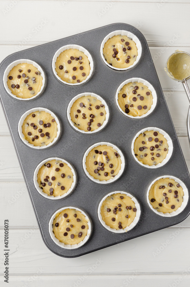 Obraz premium Raw dough for muffins with chocolate chips in baking form on a white wooden background. Recipe step by step. Top view.