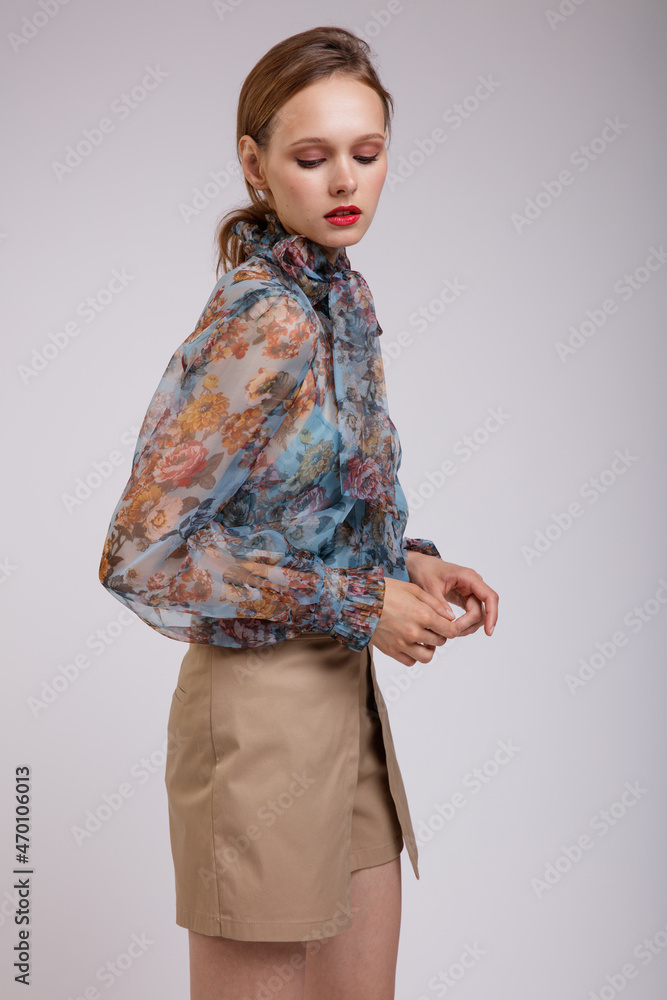 High fashion photo of a beautiful elegant young woman in a pretty blue transparent blouse with a floral pattern, a bow on the neck, short beige shorts similar to a skirt posing over white background High fashion photo of a beautiful elegant young woman in a pretty blue transparent blouse with a floral pattern, a bow on the neck, short beige shorts similar to a skirt posing over white background