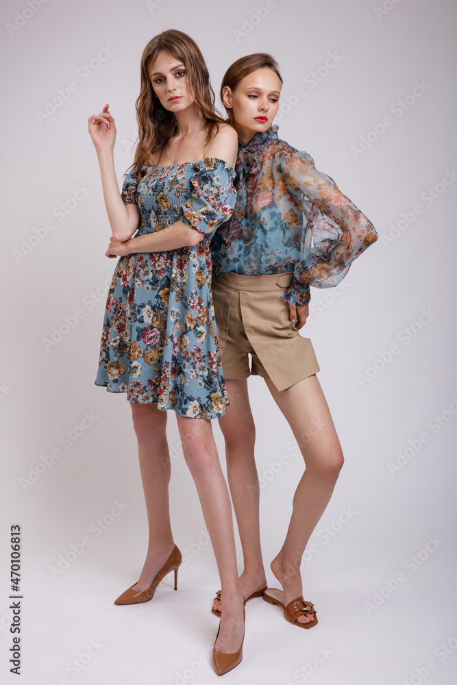 Two high fashion models in blue transparent blouse with floral pattern, dress, beige shorts. Beautiful young women. Studio shot. White background. Slim figure. Make up, hairstyle. Two high fashion models in blue transparent blouse with floral pattern, dress, beige shorts. Beautiful young women. Studio shot. White background. Slim figure. Make up, hairstyle.