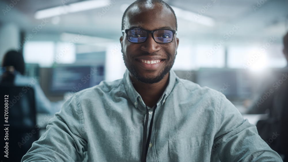 Office: Portrait of Focused IT Programmer Wearing Headphones Working on Computer, Looking at Camera and Smiling. Black Male Software Engineer Developing Innovative App, Program, Video Game. Office: Portrait of Focused IT Programmer Wearing Headphones Working on Computer, Looking at Camera and Smiling. Black Male Software Engineer Developing Innovative App, Program, Video Game.