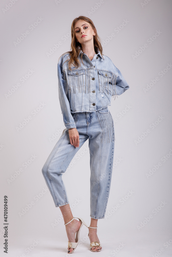 High fashion photo of a beautiful elegant young woman in a pretty denim jeans oversize jacket with rhinestones, pants posing over white, soft gray background. Slim figure. Studio Shot. High fashion photo of a beautiful elegant young woman in a pretty denim jeans oversize jacket with rhinestones, pants posing over white, soft gray background. Slim figure. Studio Shot.