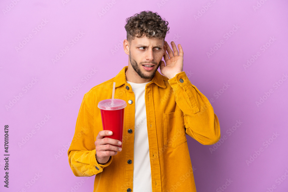 Young blonde man holding soda isolated on purple background listening to something by putting hand on the ear Young blonde man holding soda isolated on purple background listening to something by putting hand on the ear