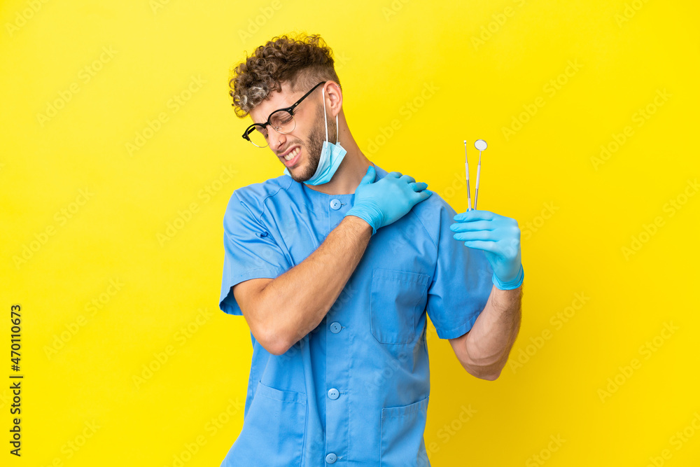Dentist blonde man holding tools isolated on background suffering from pain in shoulder for having made an effort Dentist blonde man holding tools isolated on background suffering from pain in shoulder for having made an effort