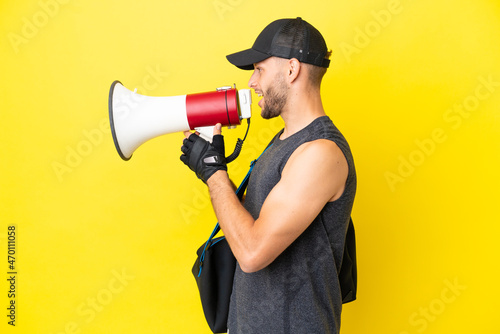 Young sport blonde man with sport bag isolated on yellow background shouting through a megaphone