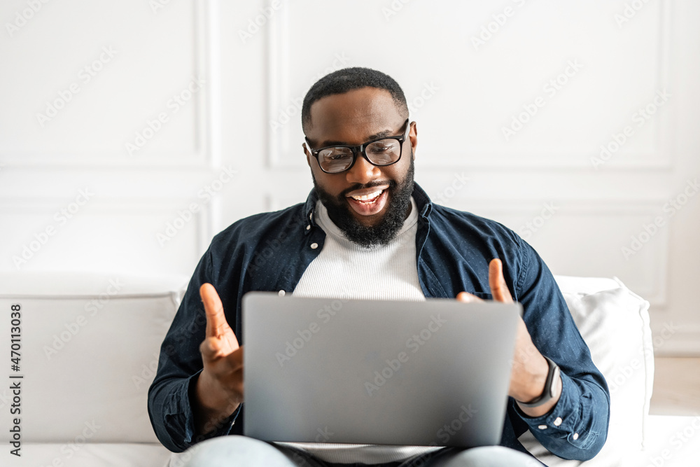 Positive African-American guy using laptop for video call sitting on the sofa at home. Cheerful black man taking a part in virtual meeting, involved virtual webinar, look at webcam and talking Positive African-American guy using laptop for video call sitting on the sofa at home. Cheerful black man taking a part in virtual meeting, involved virtual webinar, look at webcam and talking