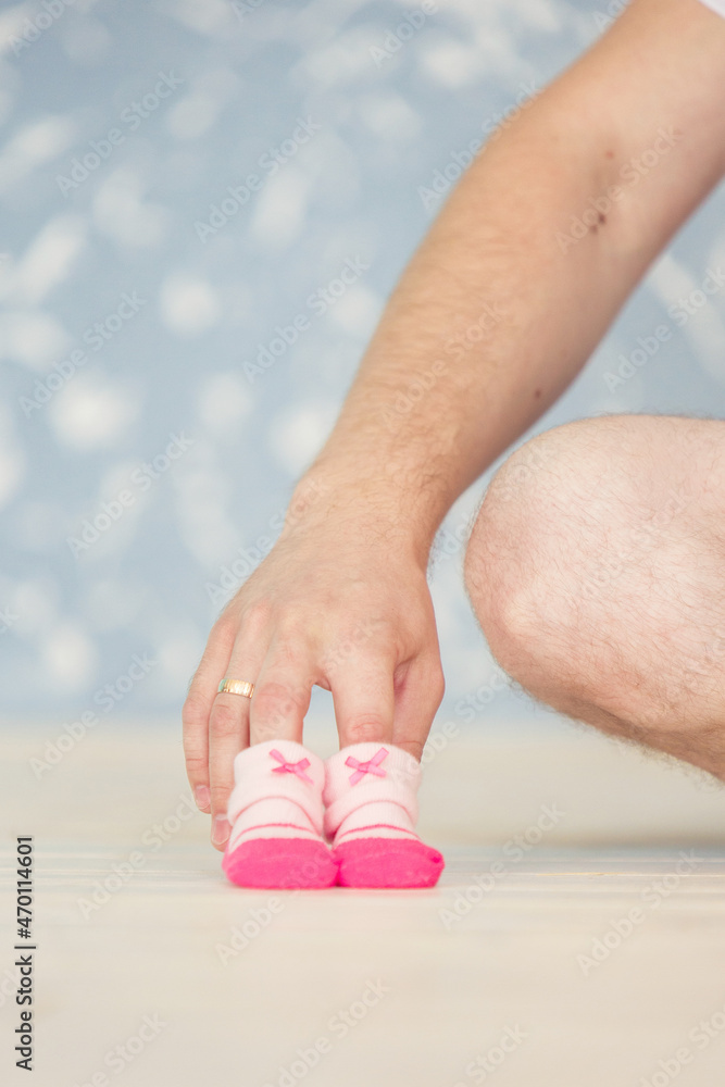 father-to-be sits on the floor against a blurred blue background and holds pink soft newborn shoes standing on the beige floor