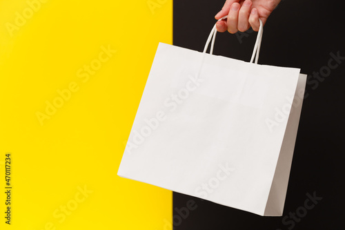 Female hand holding white blank shopping bag isolated on yellow and black background. Black friday sale, discount, recycling, shopping and ecology concept.