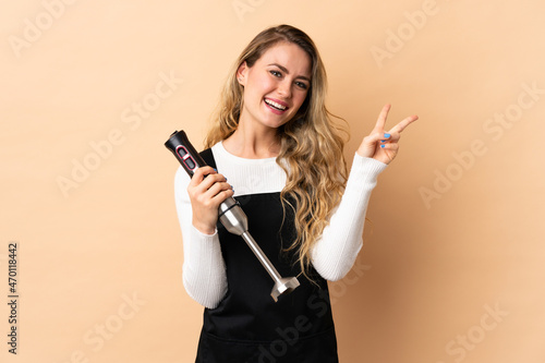 Young brazilian woman using hand blender isolated on beige background smiling and showing victory sign