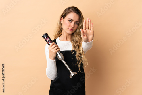 Young brazilian woman using hand blender isolated on beige background making stop gesture