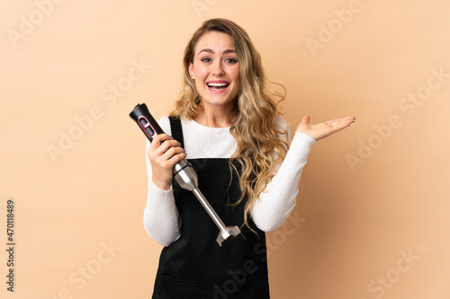 Young brazilian woman using hand blender isolated on beige background with shocked facial expression