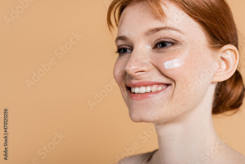 Smiling redhead woman with perfect skin and cream on face looking away isolated on beige