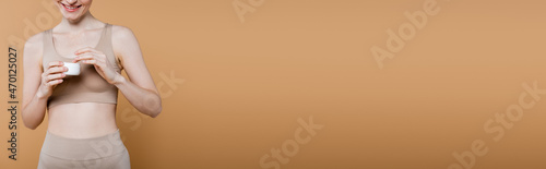 Cropped view of positive woman in top holding cosmetic cream isolated on beige, banner