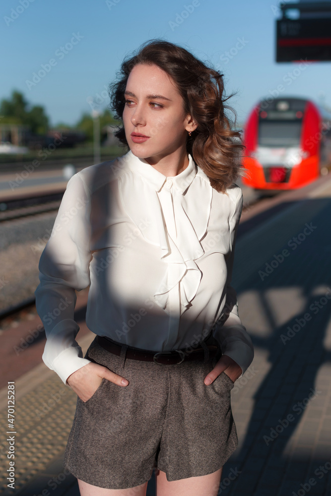 Attractive young woman, in a white shirt, stands at the railway station next to the train.