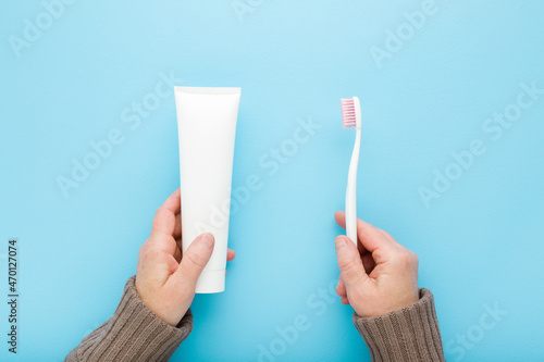 Mature woman hands holding and showing white tube of toothpaste and toothbrush on light blue table background. Pastel color. Point of view shot. Teeth hygiene in old age. Closeup. Top down view.