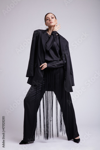 High fashion photo of a beautiful elegant young woman in a pretty black jacket, blouse, fringe trousers, accessories over gray background. Studio Shot. Gathered dark hair. Slim figure
