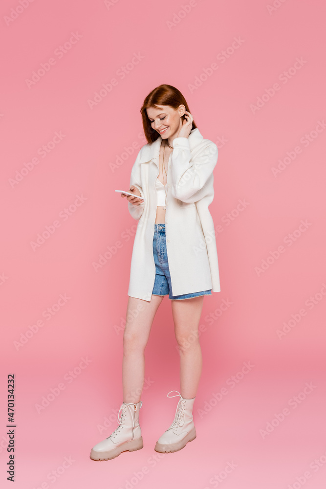 Trendy red haired woman smiling while using cellphone on pink background Trendy red haired woman smiling while using cellphone on pink background