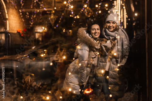two beautiful young women smiling and hugging, reflected in a showcase with Christmas lights.