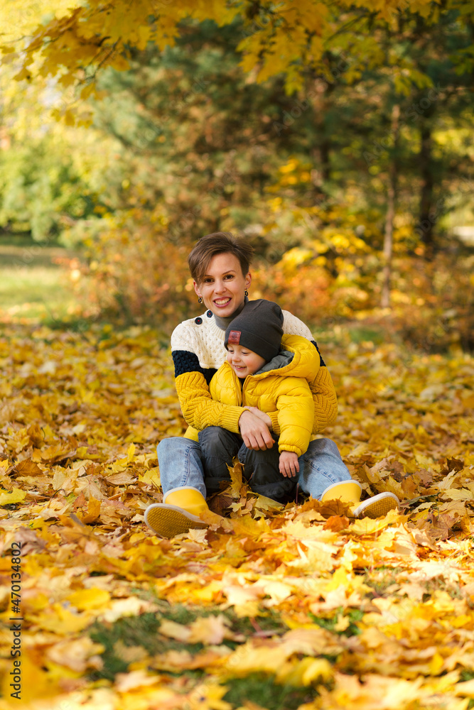 Happy young mother hugging her little son and sitting in autumn park among the yellow leaves Happy young mother hugging her little son and sitting in autumn park among the yellow leaves