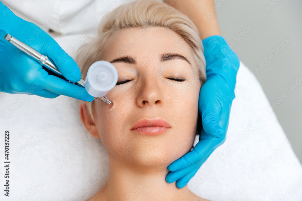 Naklejka premium Cosmetologist in blue gloves does a gas-liquid peeling procedure of the client's face. Top view. The concept of professional cosmetology