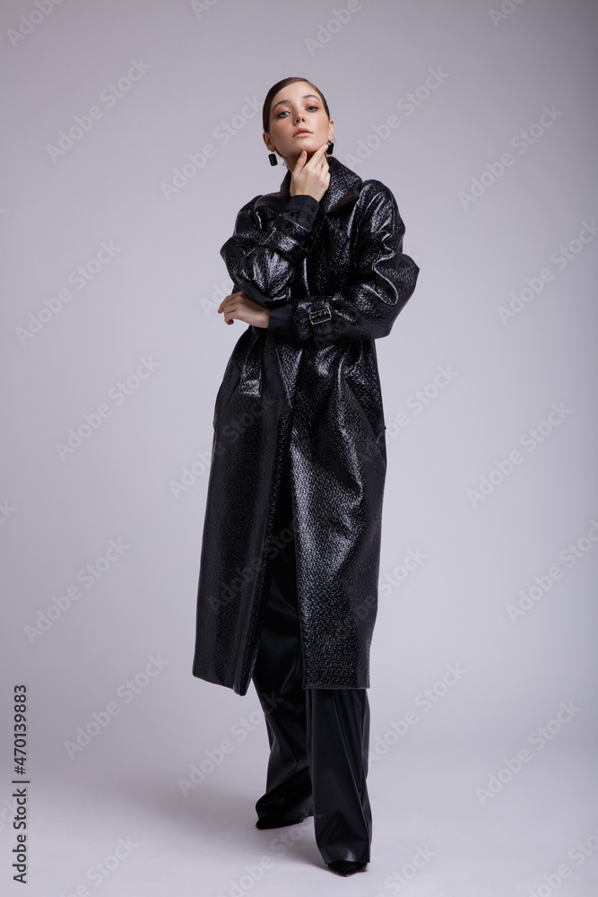 High fashion photo of a beautiful elegant young woman in a pretty leather coat, pants,  accessories posing over white, soft gray background. Studio Shot. Gathered dark hair. Slim figure