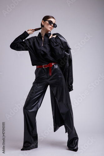 High fashion photo of a beautiful elegant young woman in a pretty leather coat, pants, stylish sunglasses, accessories, shirt, red belt posing over gray background. Studio Shot. Gathered dark hair