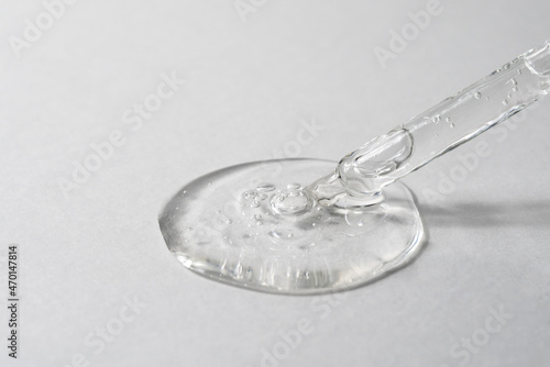 Facial serum pouring from glass pipette on grey textured background. Transparent liquid gel drop with bubbles closeup. Skin care routine. Trendy cosmetic product macro. Front view. © gorina_anna