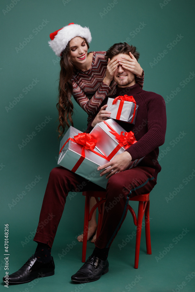Fototapeta premium Beautiful young woman, evening makeup stylish dress , brunette, and young man in sweater, gift boxes red silk bows New Year, Christmas. Fashion photo. Green background. New year's Christmas cap
