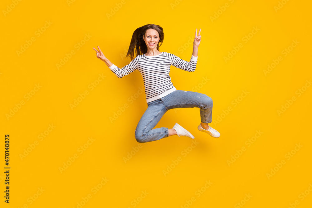Full size photo of lovely brunette millennial lady jump show v-sign wear sweater jeans sneakers isolated on yellow color background Full size photo of lovely brunette millennial lady jump show v-sign wear sweater jeans sneakers isolated on yellow color background