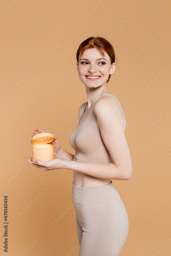 Cheerful woman with perfect skin holding container with cosmetic cream isolated on beige