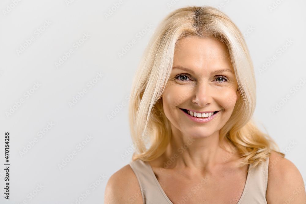 Fototapeta premium Headshot of gorgeous beautiful middle-aged woman with pure and healthy skin and blonde hair, stands isolated on grey background, mature lady looks at camera and smiles, copy space