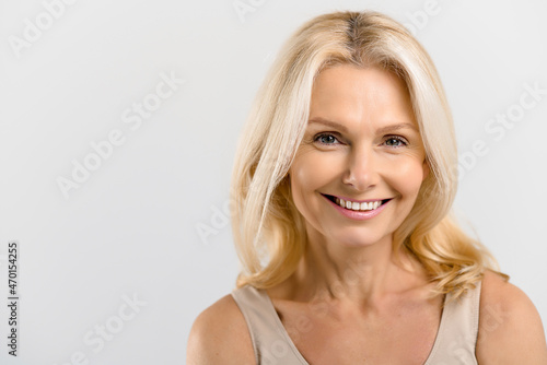 Headshot of gorgeous beautiful middle-aged woman with pure and healthy skin and blonde hair, stands isolated on grey background, mature lady looks at camera and smiles, copy space