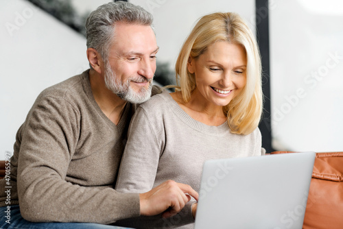 Lovely mature spouses using laptop computer for spending leisure at home, watching movie together, choosing purchases, shopping. Middle-aged couple sits on the sofa and watching at the screen