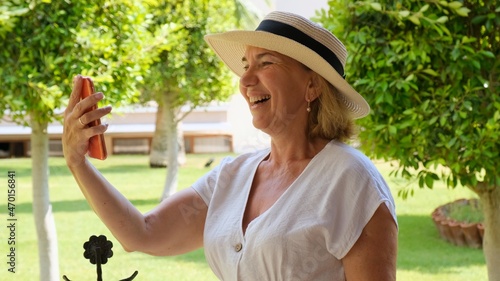 blonde senora female, 50-55 years old, in a hat and a white dress, talks using a smartphone via video call on her terrace on a sunny morning on a summer day. Freelance concept, woman blogger, business