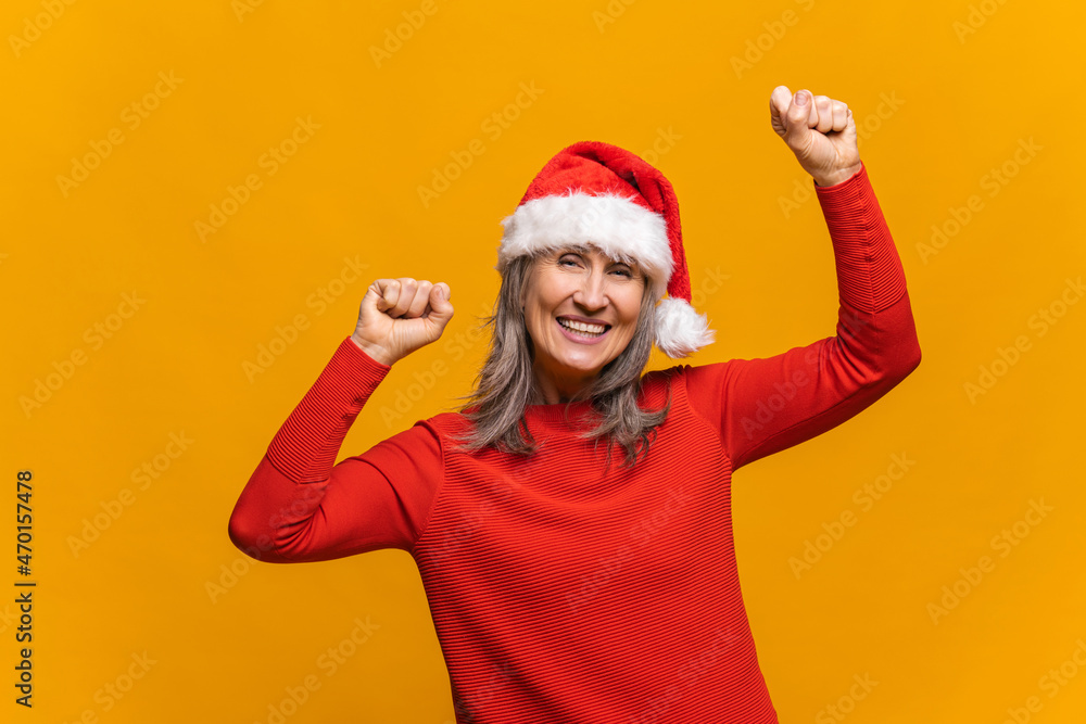 Holiday mood concept. Joyful and cheerful senior gray-haired woman in santa hat raising arms and dancing, mature lady looks at the camera with a toothy smile, celebrating new year, isolated on yellow Holiday mood concept. Joyful and cheerful senior gray-haired woman in santa hat raising arms and dancing, mature lady looks at the camera with a toothy smile, celebrating new year, isolated on yellow
