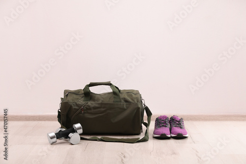 Green sports bag, sneakers and dumbbells on floor near white wall, space for text