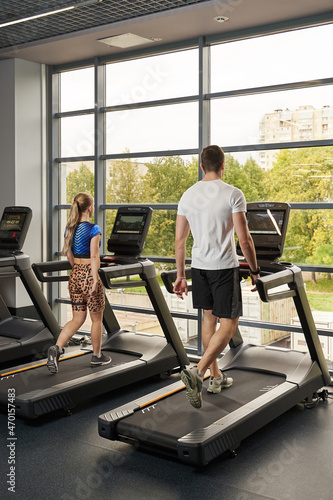 a man and a woman walk on a treadmill simulator in the gym in front of a huge window. back view
