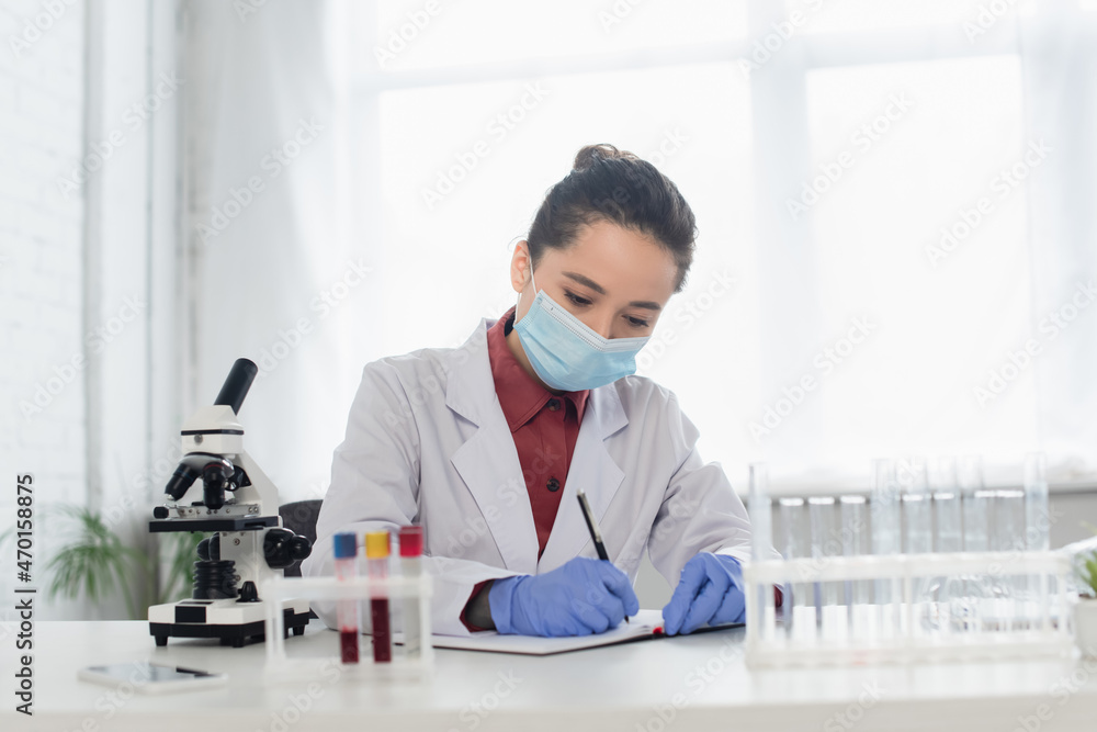 scientist in latex gloves and medical mask writing in notebook near test tubes and microscope. scientist in latex gloves and medical mask writing in notebook near test tubes and microscope.
